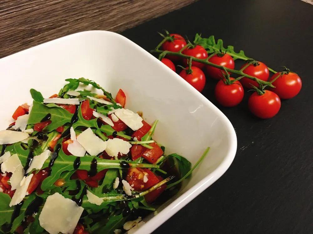 a salad in a white bowl with tomatoes and parmesan cheese on the side